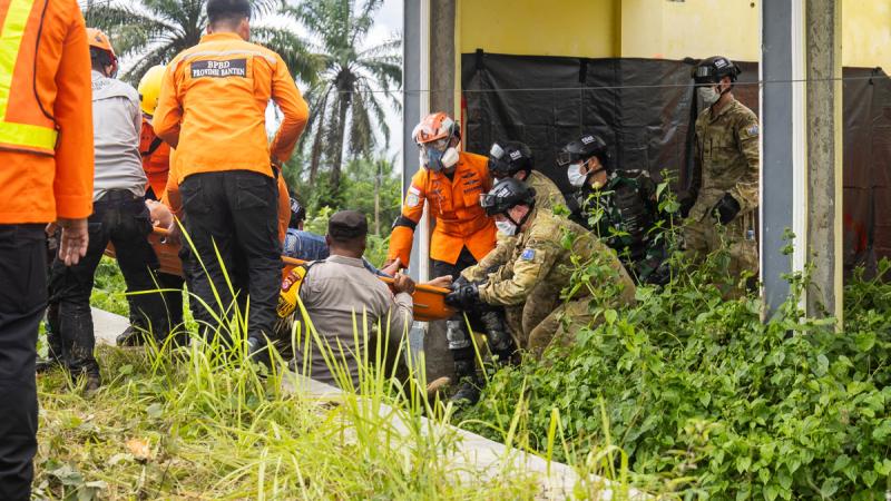 A group of personnel, many wearing helmets and medical masks, manoeuvre someone on a stretcher.