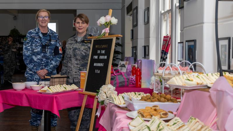 Two women in military uniform stand beside a pink display with a variety of snacks.