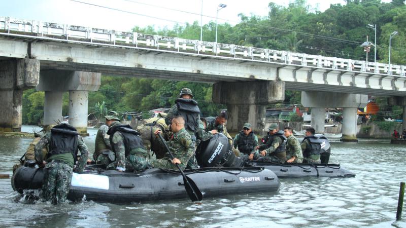 Two inflatable boats carrying Australian soldiers and Philippine marines float down a river near a concrete bridge, with one marine using a paddle.