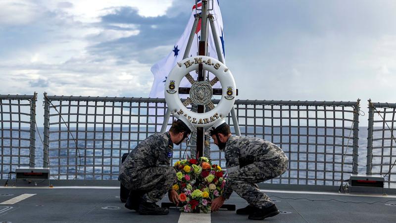 Two sailors crouch to lay a wreath in front of a flagpole on a ship.