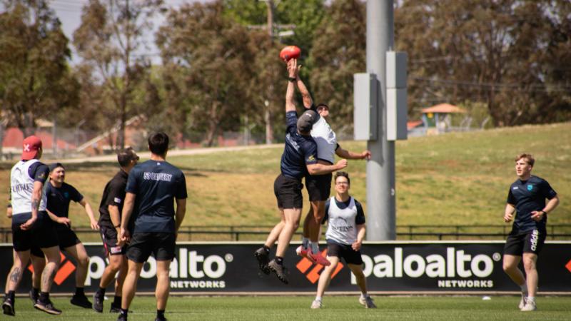 Two men leaping very high, both reaching for the AFL ball as their teammates look on.