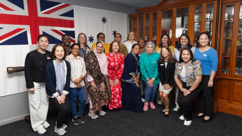 A group of women pose for a photo indoors in front of a large Royal Australian Navy ensign and a display cabinet.