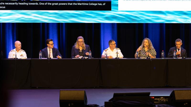 Six panellists seated on stage with a large screen behind them.