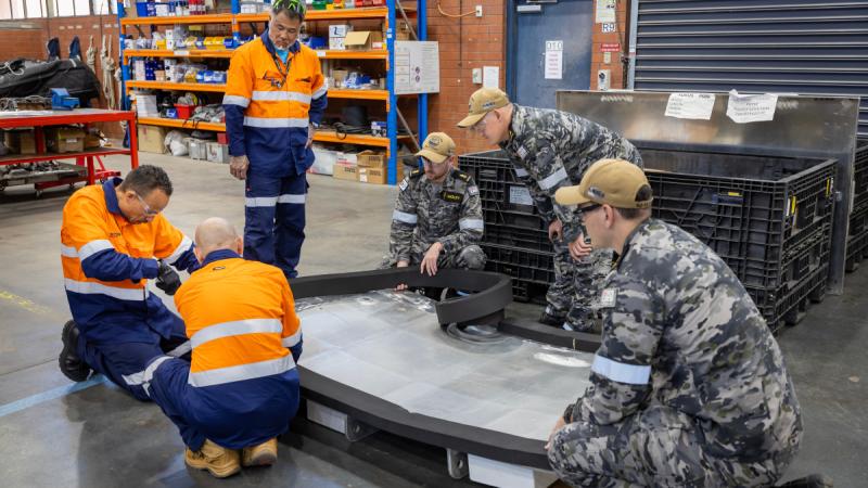 Several technicians, some in high-vis clothing, are crouched around a large curved panel, attaching thick black rubber to it, in a spacious workshop with tools and equipment visible in the background.