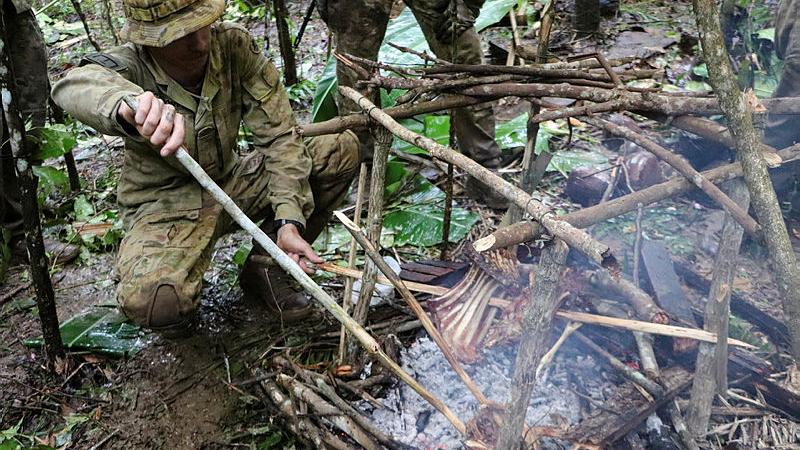 A man crouches next to a smoking fire and pokes it with a stick while animal ribs are cooking on a spit.