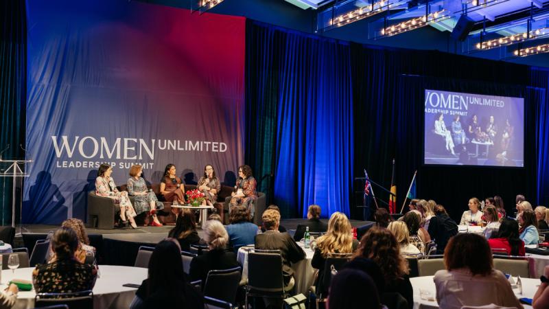Women lead a discussion from armchairs on stage while attendees listen from tables in a spacious event hall.