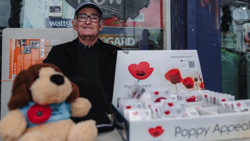 An elderly man sits at a stall outside a shop with ‘Poppy Appeal’ poppies and a stuffed dog wearing a poppy, offering poppies for sale.