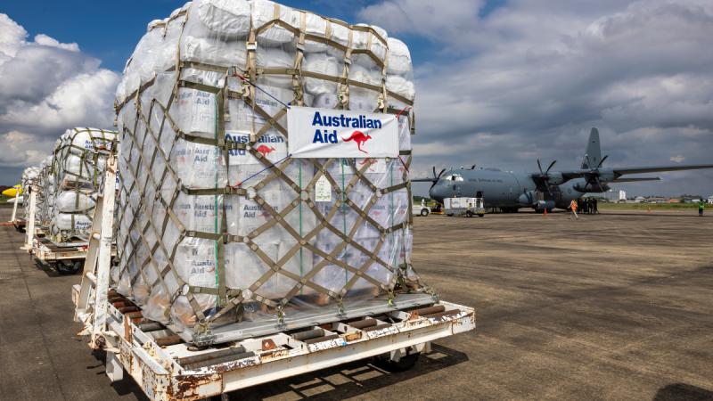 Australian Aid pallets are transported across a tarmac in the foreground, with a C-130J Hercules in the background.