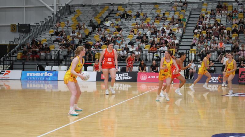 Netball players in yellow and red uniforms focus on the ball during a game.
