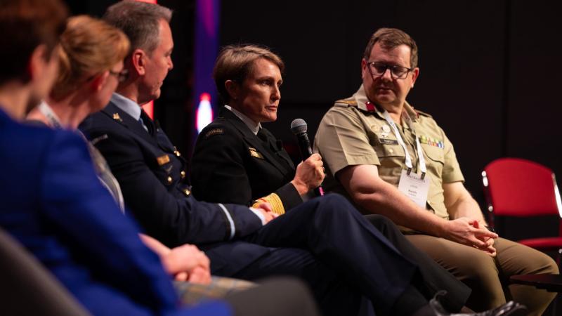 A senior naval officer seaks at a conference, seated with colleagues.
