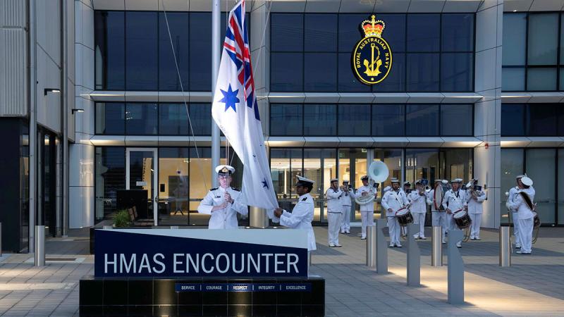 Navy personnel in white uniforms stand in formation near a white flag and in front of a building.