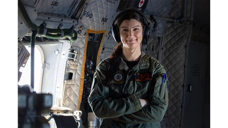 A woman in military uniform and headset stands inside an aircraft.