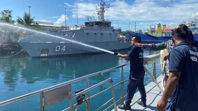 One maritime police member holds and points a hose out to the ocean where a ship is seen in the background on a fine, partly cloudy day.