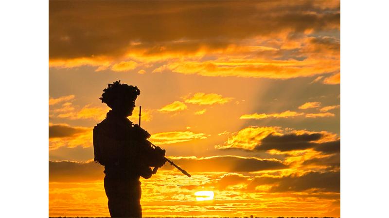An armed military member stands before a sunset.