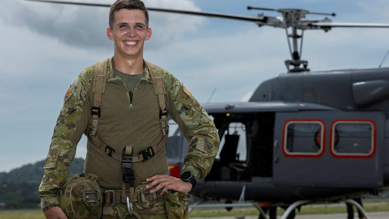 A soldier stands with his helmet in one hand and his other hand on his hip in front of an Army helicopter.