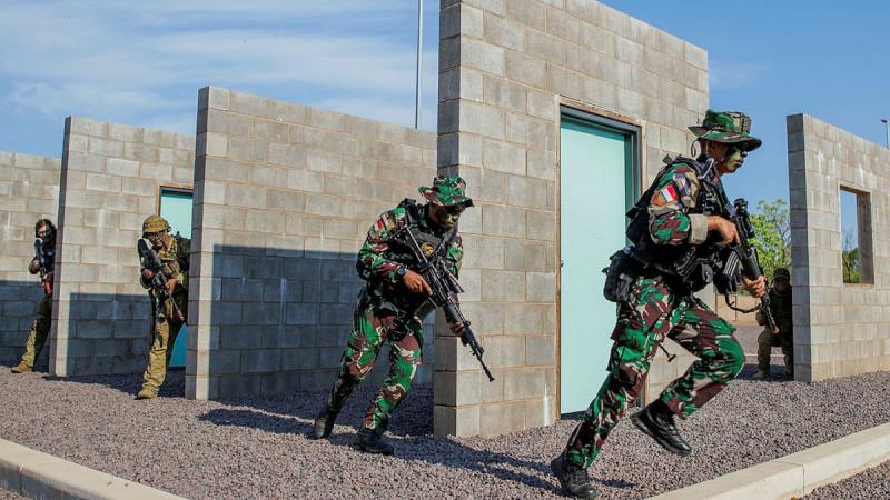 Four soldiers partially hide and then run out from behind concrete walls with guns in hand.