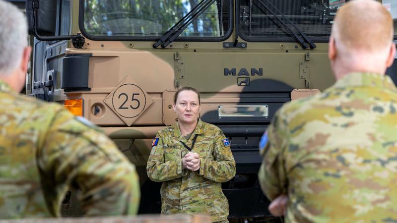 A female Army member stands with her arms crossed - facing two other Army members - in front of an Army vehicle.