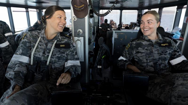 Two women in Navy uniforms sit side by side and smile at each other inside a ship.