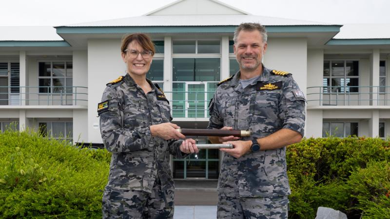 Two Navy officers stand side by side outside a 2-storey building, exchanging a nautical telescope and smiling.