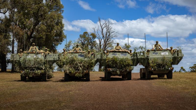Four high mobility artillery rocket system vehicles, each with three soldiers on them, camouflaged with tree branches on the front, parked in a straight row.