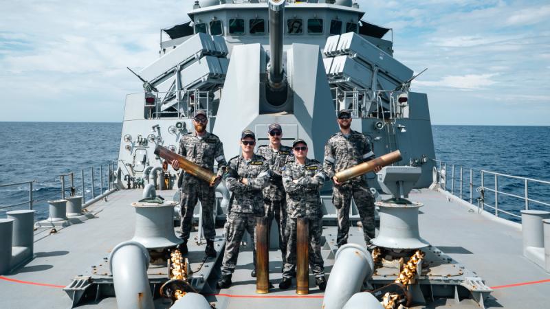 Five men stand on a ship’s bridge beneath a large gun, two holding shells with two more shells upright on the deck. 