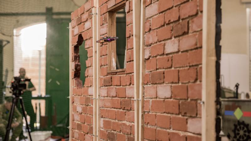 A drone hovers in front of a window opening in a temporary brick wall inside a large room.