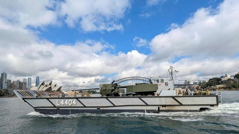 A boat carries an Army rocket system across the water on a partly cloudy day.