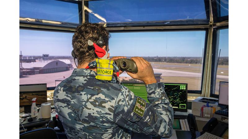 A military member looks out a window with binoculars and an elf on his shoulder.