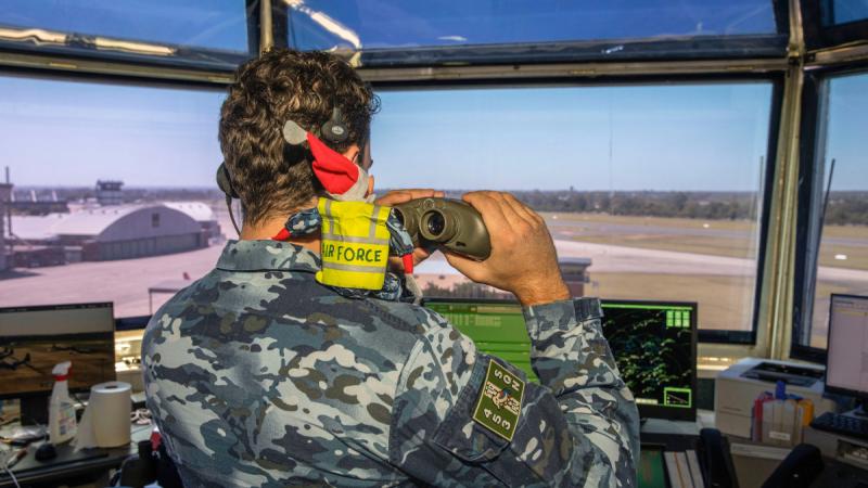 A military member looks out a window with binoculars and an elf on his shoulder.