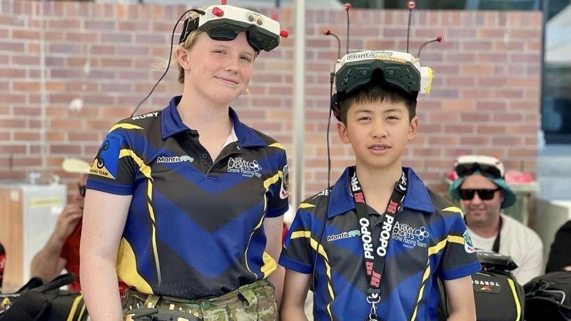 Two young Army cadets stand with drone controls in their hands and wear headgear involved in the control of the drones.