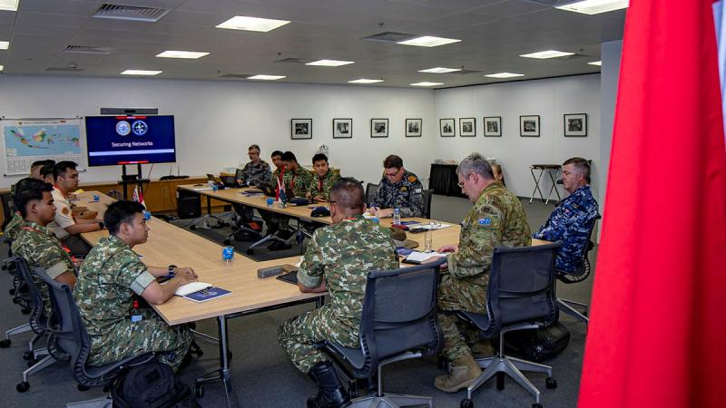 A group of military people gather in a boardroom centred around a screen.