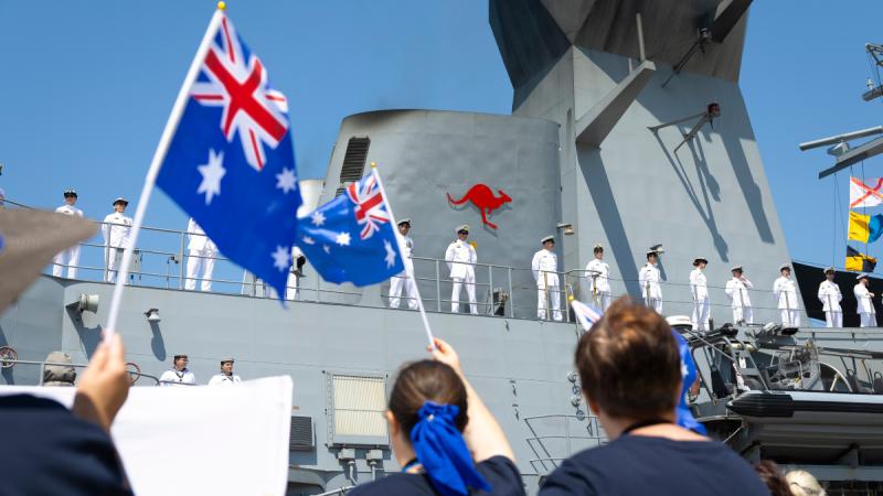 People on a quay waving Australian flags toward sailors standing on the decks of a ship.