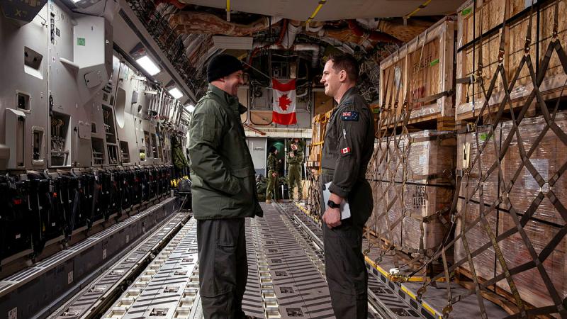 One man in uniform talks to another with crates of cargo in the background inside an aircraft.