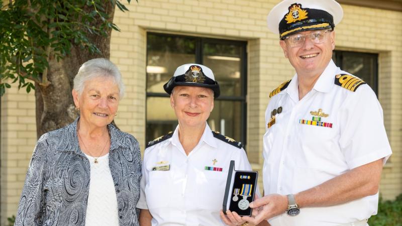 A senior Navy officer presents a medal to a junior officer, with a civilian in attendance