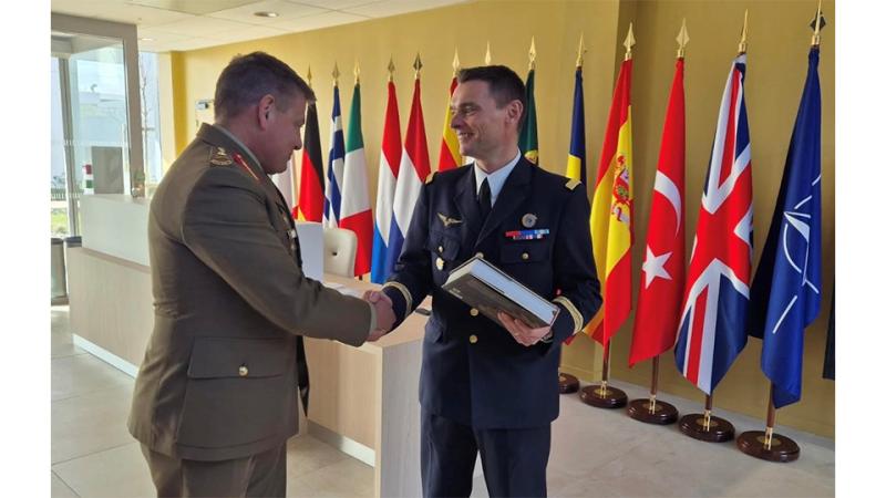 Two men in military regalia shake hands before international flags.
