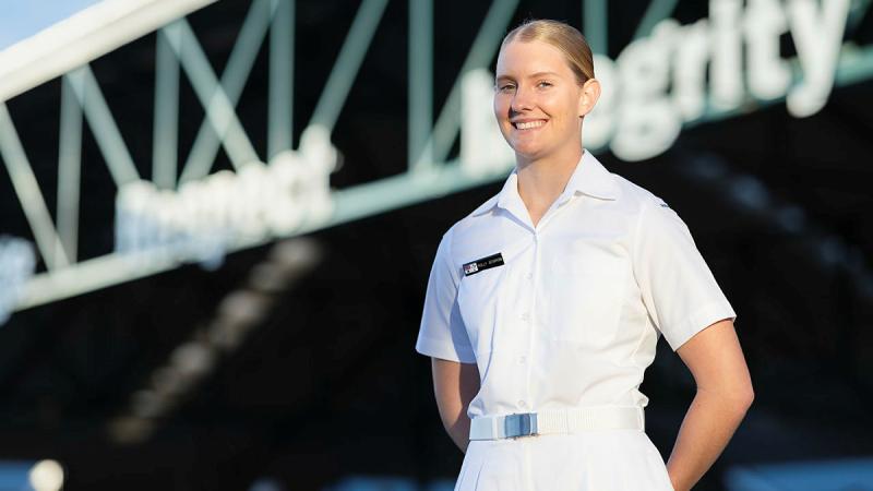 A female sailor in her Navy whites stands in front of a bridge. 