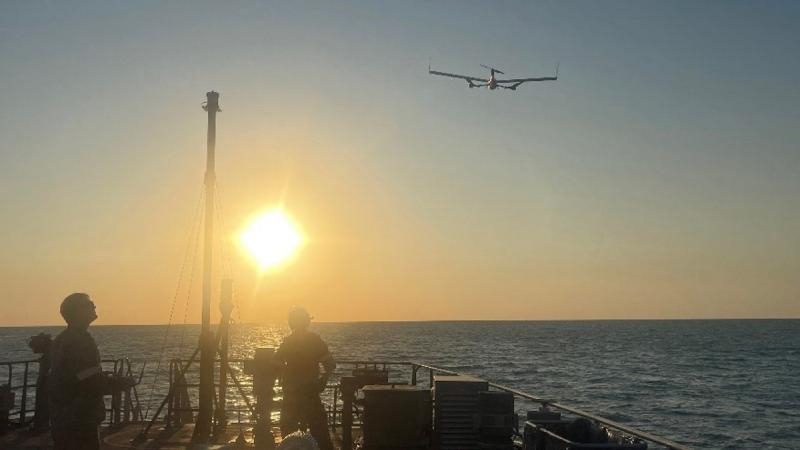 A small drone hovers above a ship’s deck as two silhouetted men stand against an orange sky, one holding a controller.
