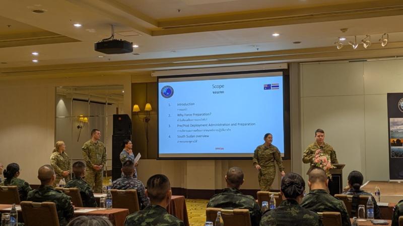 Uniformed personnel present training using a projector screen to others seated at tables in a large function room.