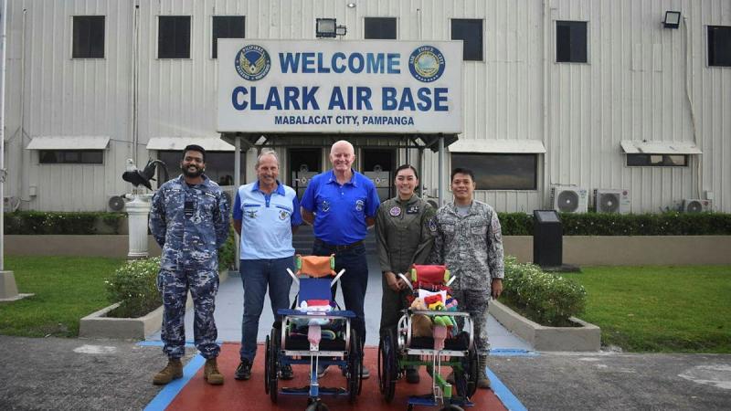 Five people with two wheelchairs stand in front of a sign that reads: Clark Air Base.