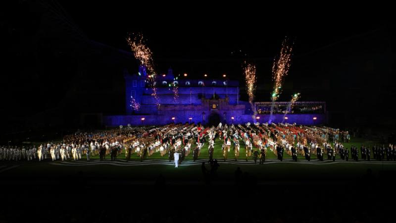 Performers fill a stadium field with a castle style stage backdrop and fireworks overhead.