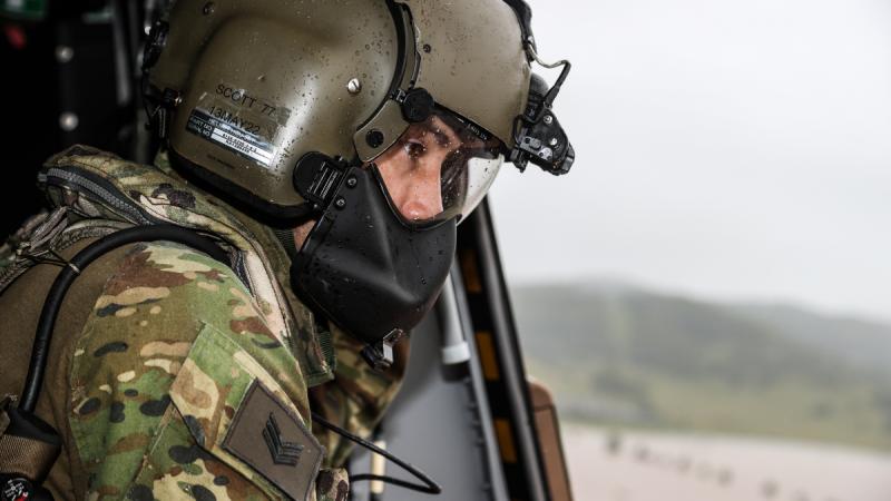 A helmeted soldier with water droplets on his gear looks out an open helicopter door toward the floodwater below.