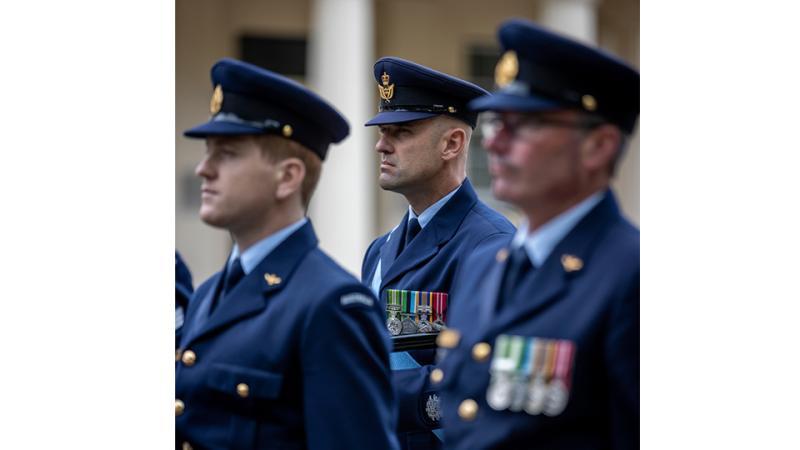 A man in ceremonial uniform stands on parade with two men blurred in the foreground.