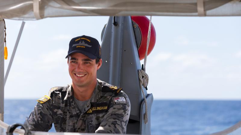 A sailor in uniform stands at a ship console with the ocean behind him.