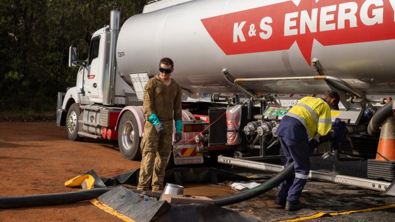 One man in high-vis attaches a hose to a petrol tanker while a soldier stands nearby.