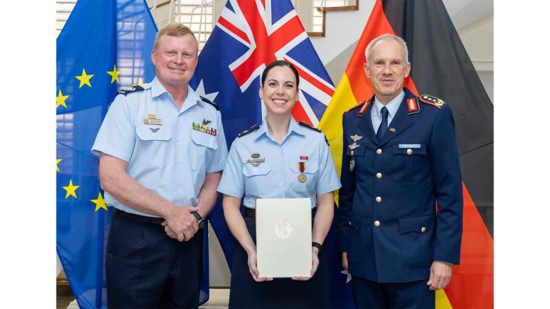 Three military personnel, one with an award, stand in front of the European flag, the Australian flag and the German flag.