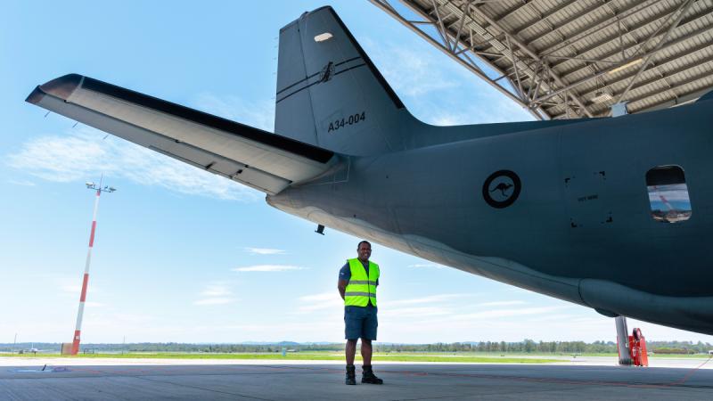 A man in a high vis vest stands near the tail of an aircraft on the flight line.