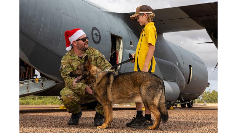 A military member with a service dog speaks to a child in front of an aircraft.