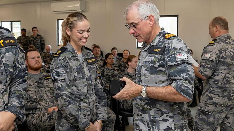 A male officer presents a female sailor with a medal in front of an audience of other sailors.