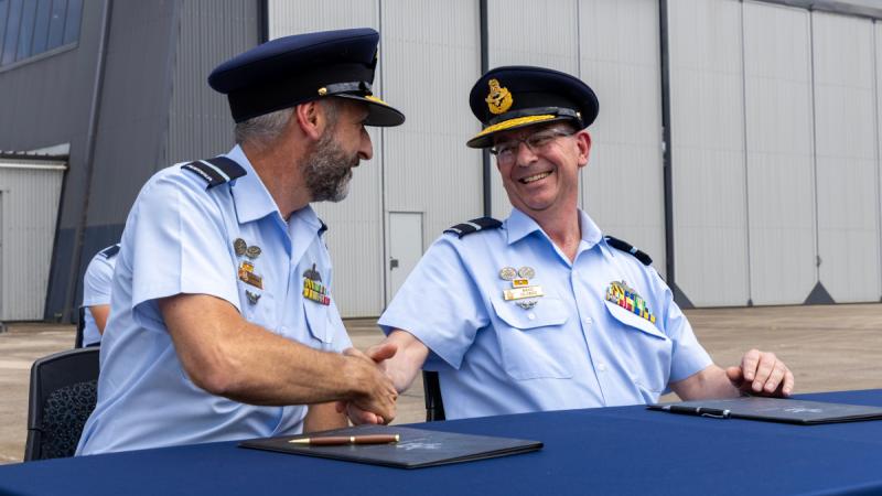 Two men in uniform sit side by side at a table on the tarmac, shaking hands with folders and pens in front of them.