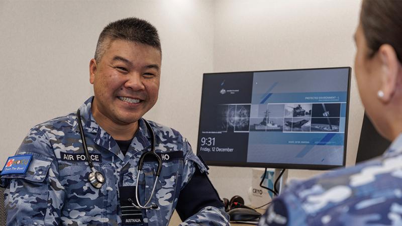 A man in Air Force fatigues with a stethoscope sits with his elbow resting on a desk, a monitor behind him and a woman opposite him.